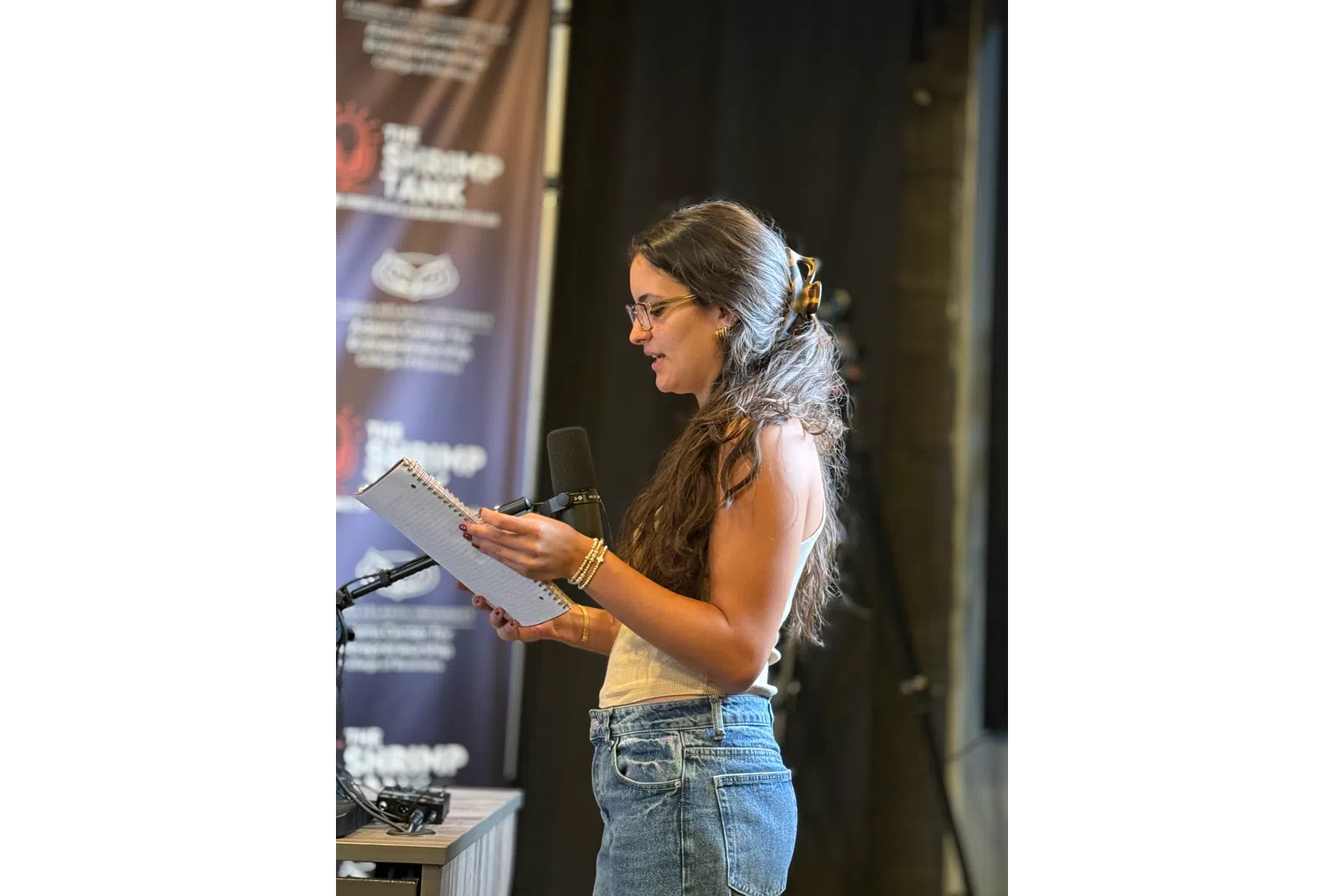 Young woman with glasses reading from a notebook into a microphone in a recording studio with banners.