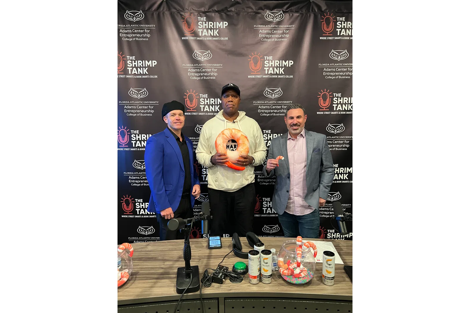 Three men standing at The Shrimp Tank event with shrimp-themed products and microphone on table