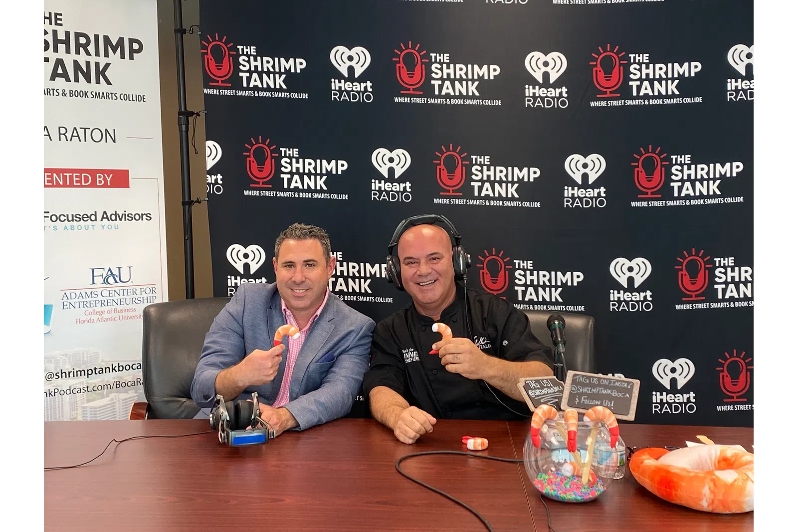 a couple of men sitting at a table with a microphone and a sign behind them