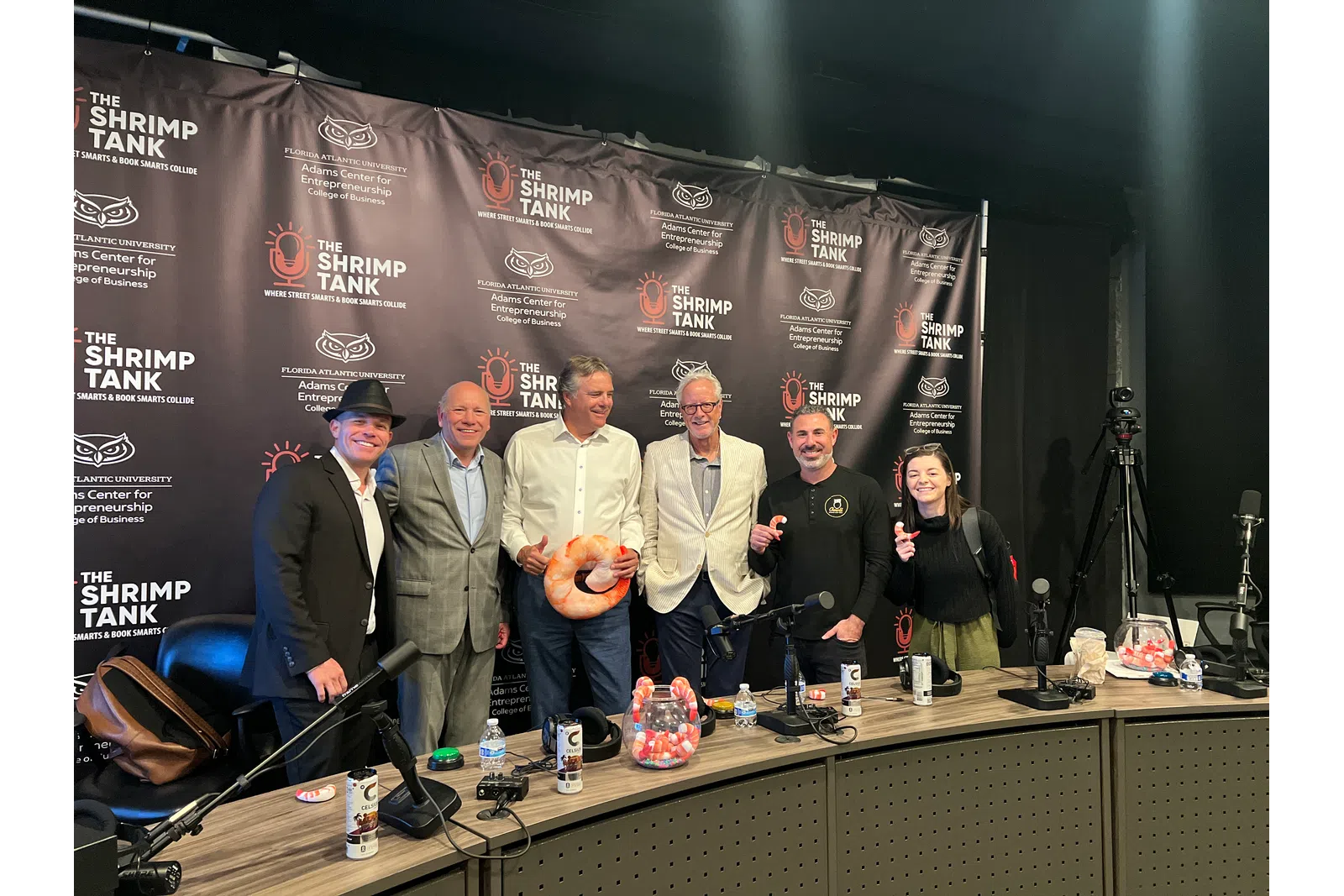Group of six people posing in front of The Shrimp Tank podcast banner in a recording studio with microphones and props.