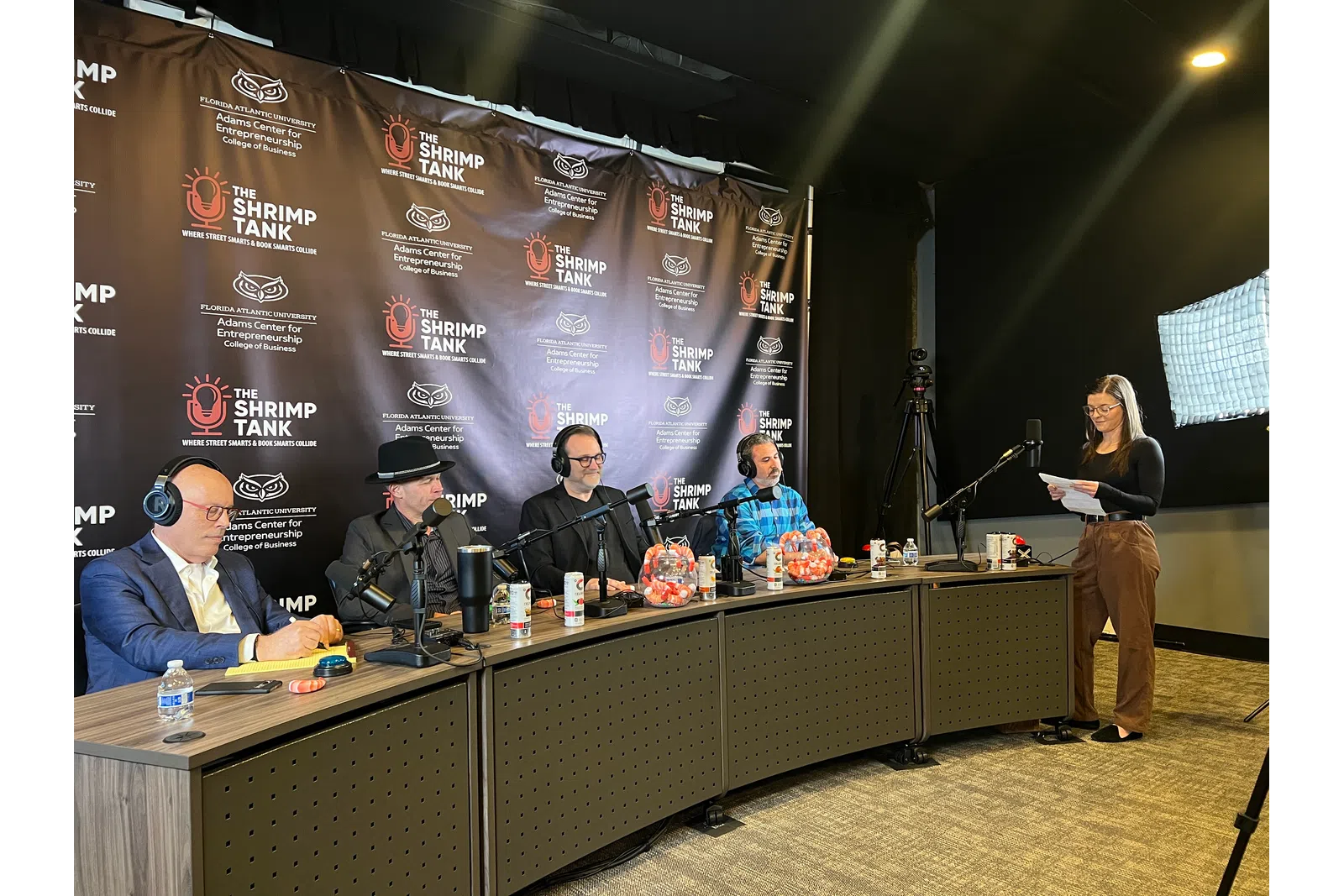 Four judges seated at a panel with microphones and headsets, a woman standing with notes in a podcast studio setup.