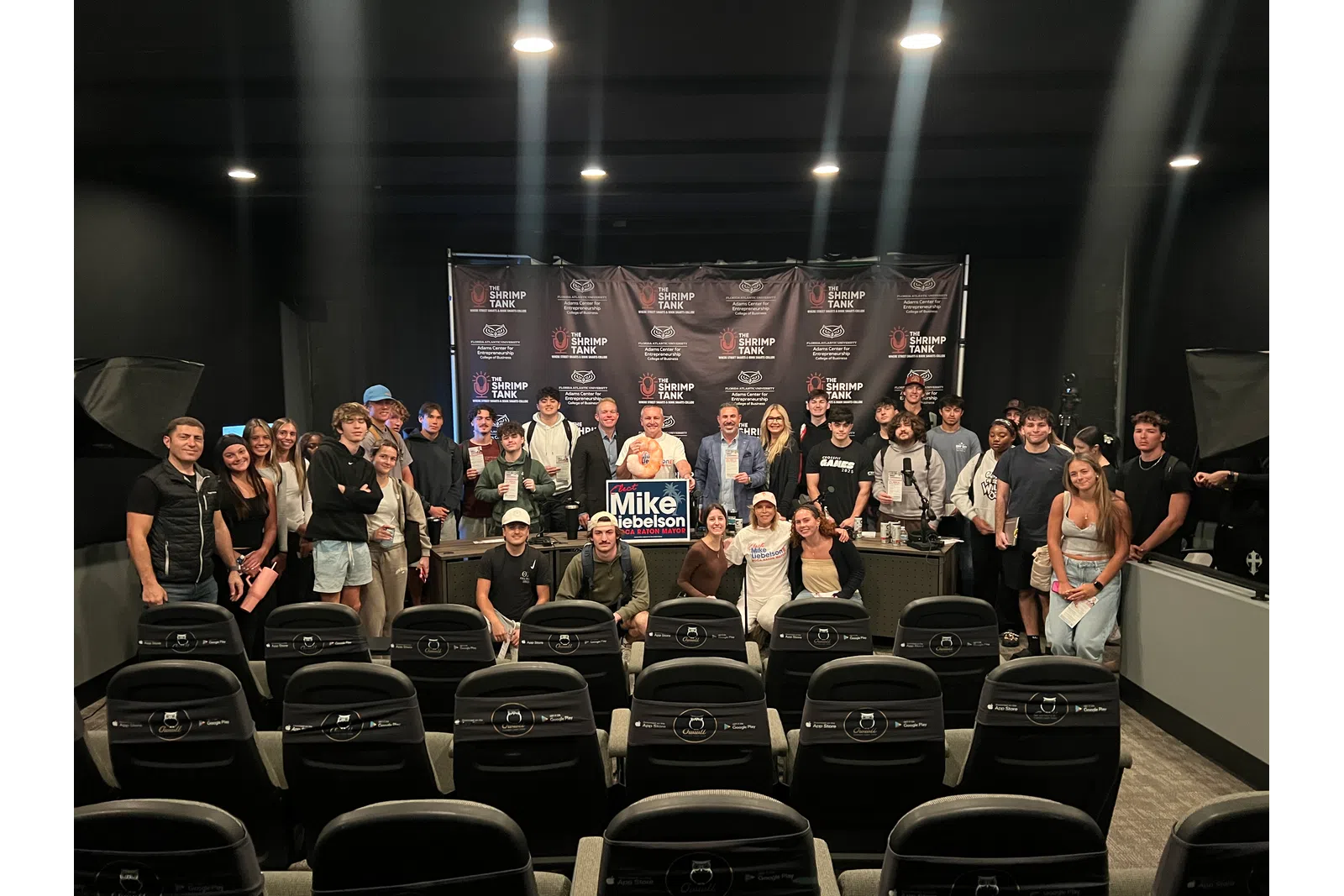 Large group of people posing for a photo in a black room with theater seats and a Mike Nebelson campaign sign