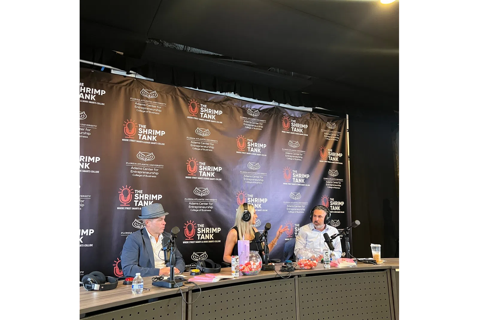 Three people recording a podcast at The Shrimp Tank event with microphones and branded backdrop.