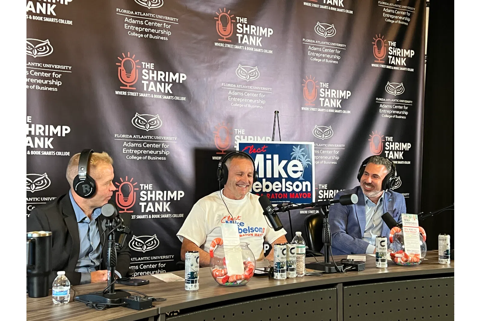Three men podcasting at The Shrimp Tank with campaign signs and branded backdrop for Mike Debelson for mayor.