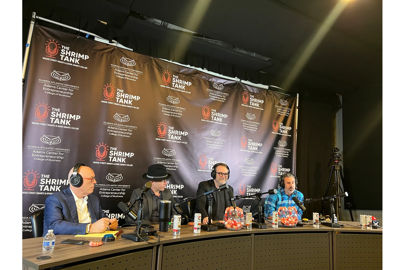Four men with headsets sitting at a podcast table with microphones and The Shrimp Tank backdrop