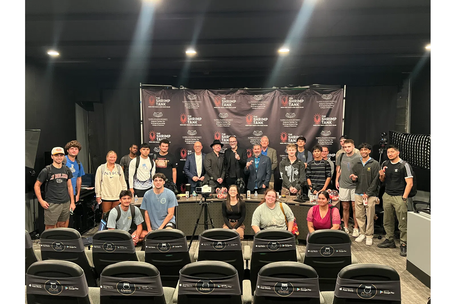 Group photo of diverse participants posing at The Shrimp Tank event with branded backdrop and professional lighting.