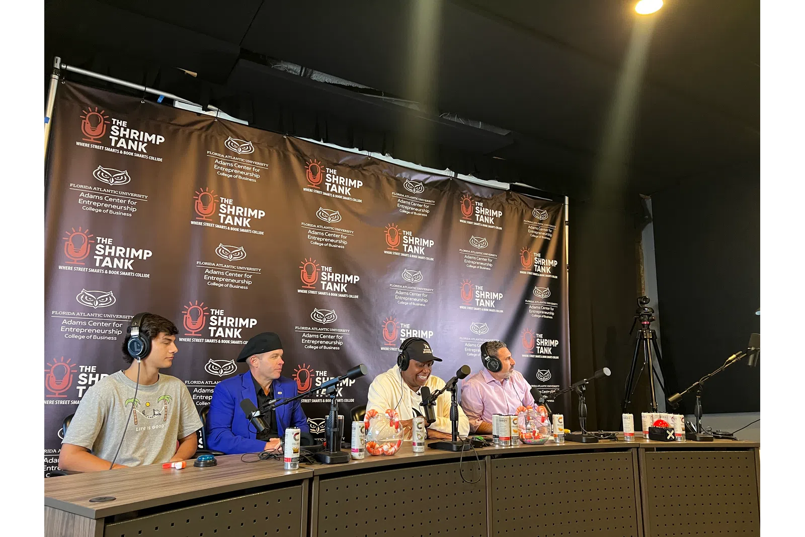 Four men with headphones sitting at a podcast panel table with microphones and branded backdrop of The Shrimp Tank event