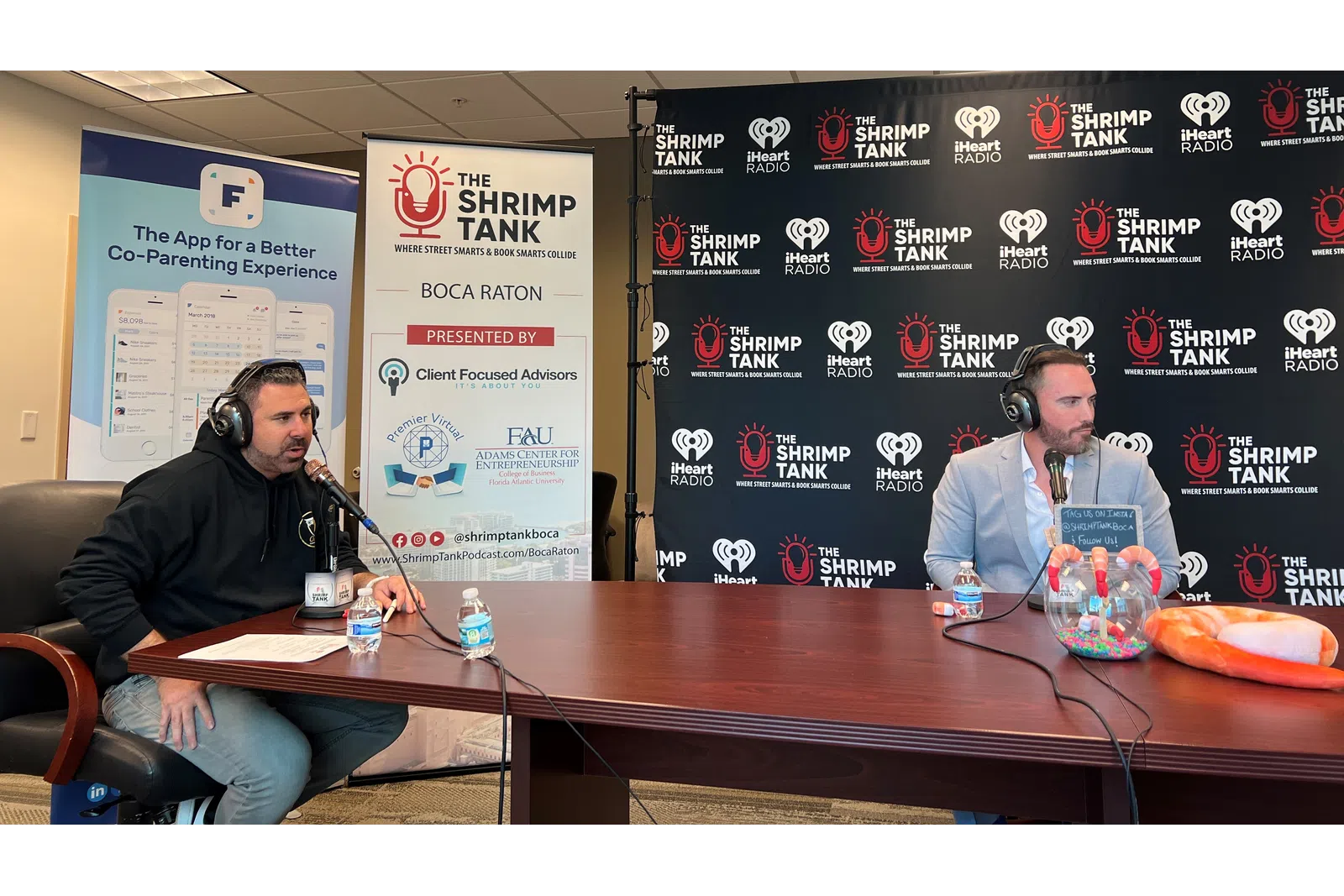 a couple of men sitting at a table with microphones and a sign