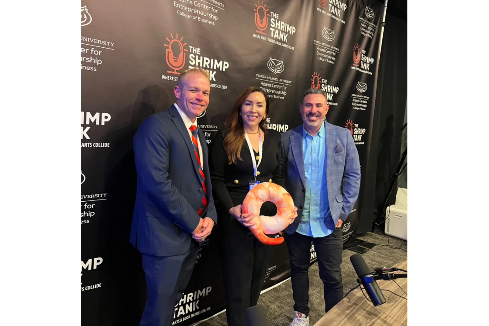 Three professionals posing at The Shrimp Tank event with branded backdrop and shrimp-shaped award