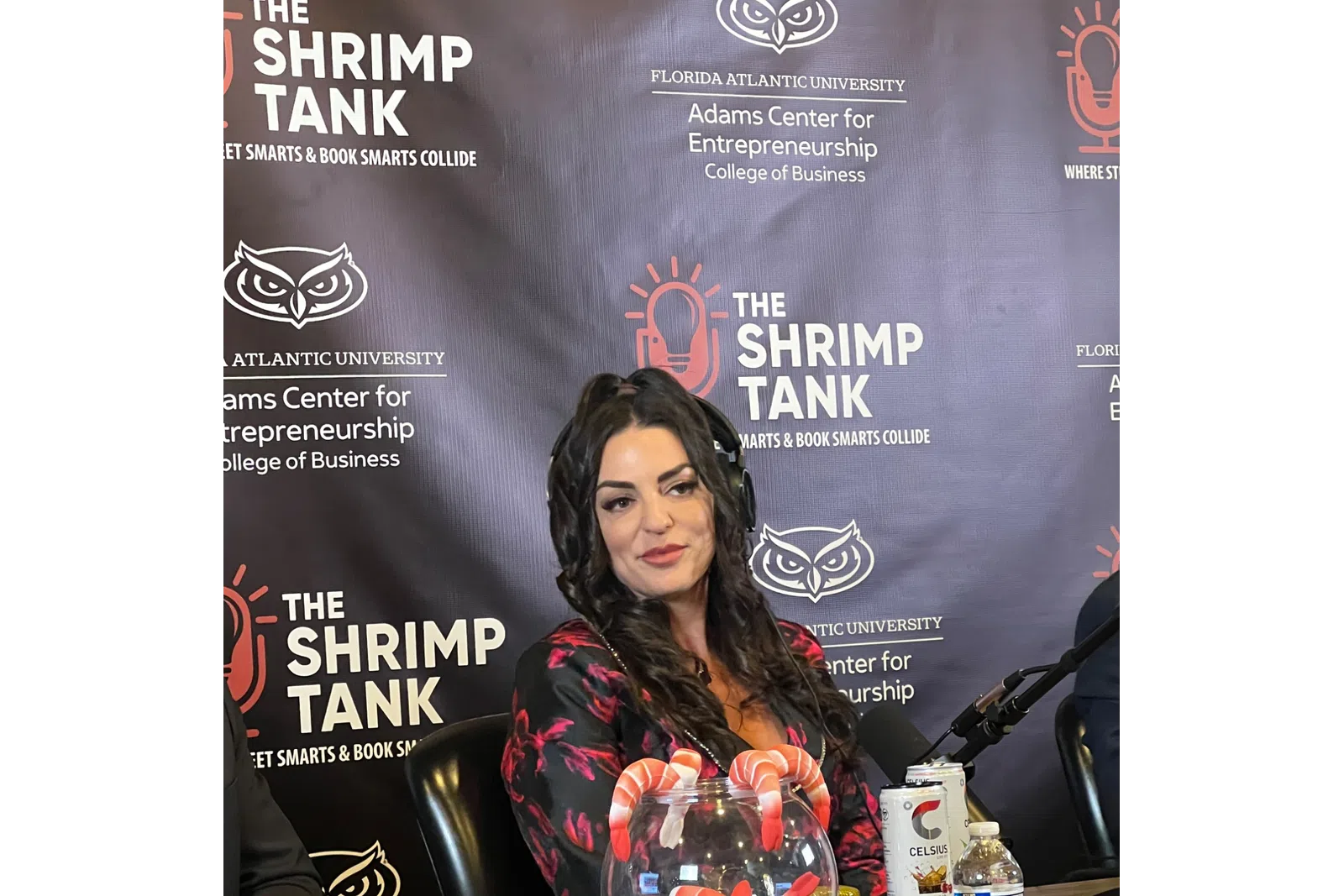 Woman sitting at desk with bowl of shrimp, microphone, and branded backdrop of The Shrimp Tank entrepreneurship event.