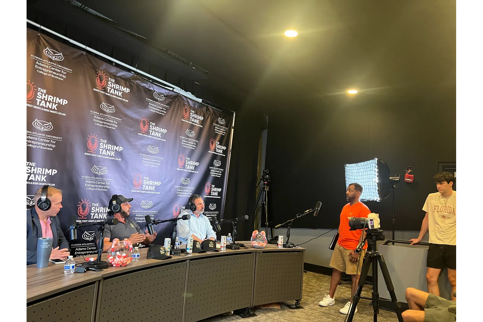 Podcast recording with four hosts behind microphones and two people standing near a camera and light setup.
