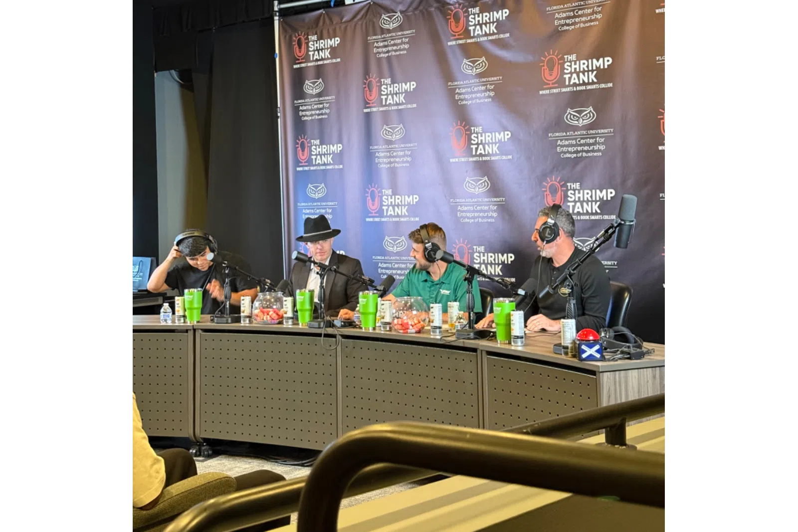 Four people wearing headphones at a podcast table with The Shrimp Tank banner and microphones