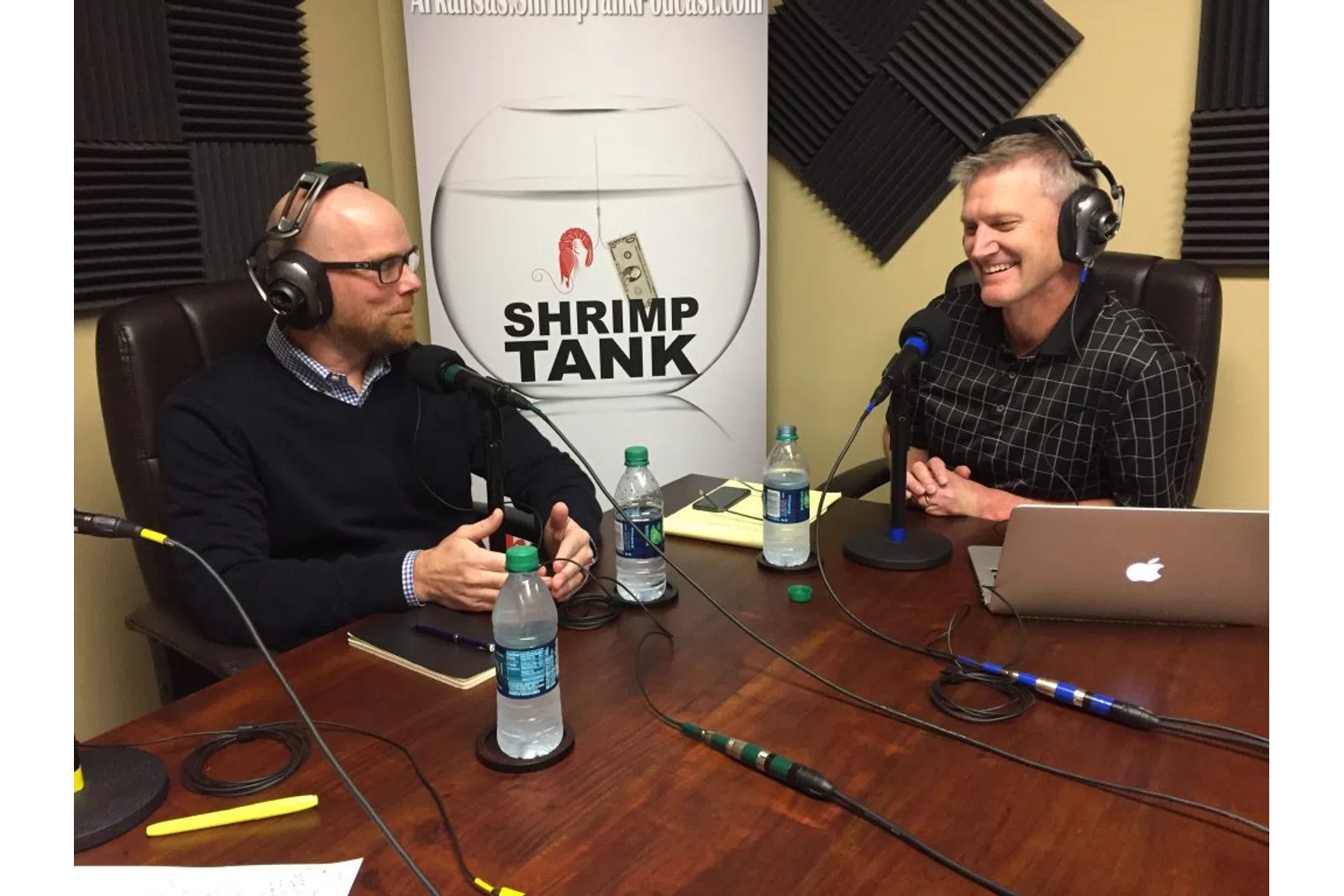a couple of men wearing headphones and sitting at a table with a laptop and microphones