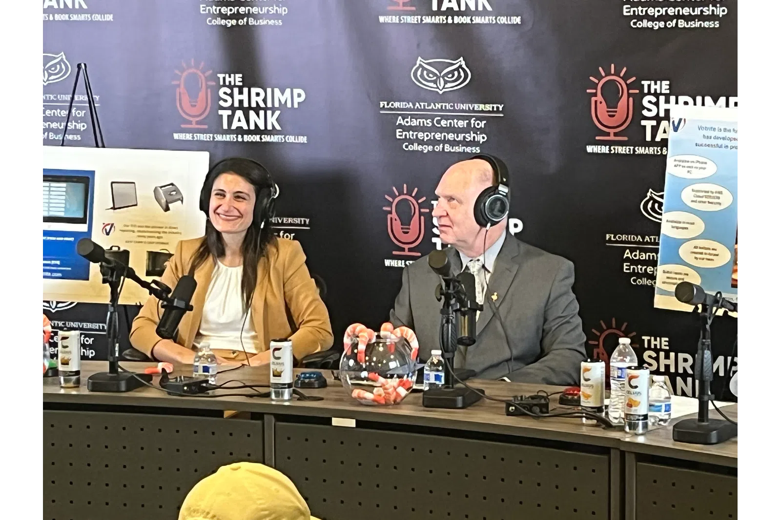 Two people with headphones speak into microphones hosting The Shrimp Tank podcast at Florida Atlantic University event