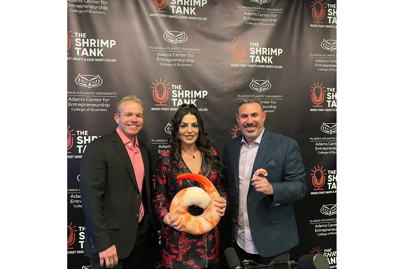 Three people pose smiling in front of a Shrimp Tank branded backdrop, one holding a large shrimp-shaped item.