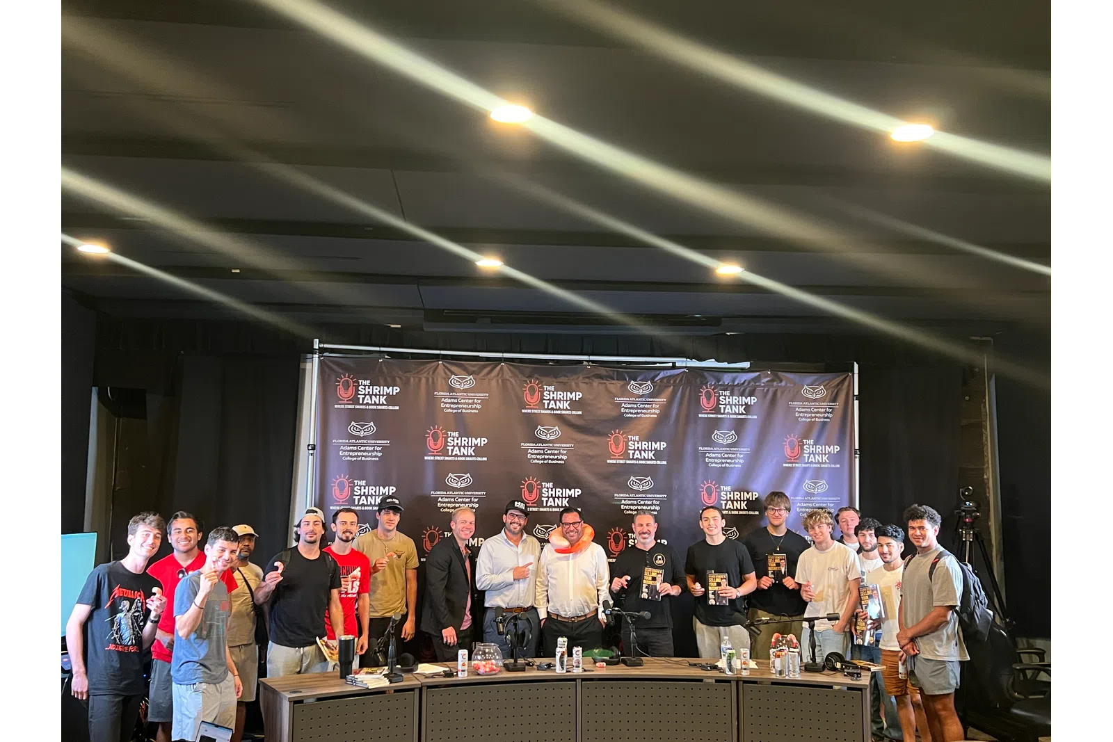 Group photo of smiling people on stage at The Shrimp Tank event with backdrop and trophies.
