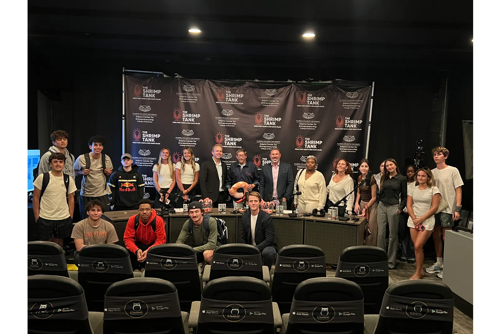 Group photo of diverse people standing and sitting in front of The Shrimp Tank branded backdrop in an event setting.