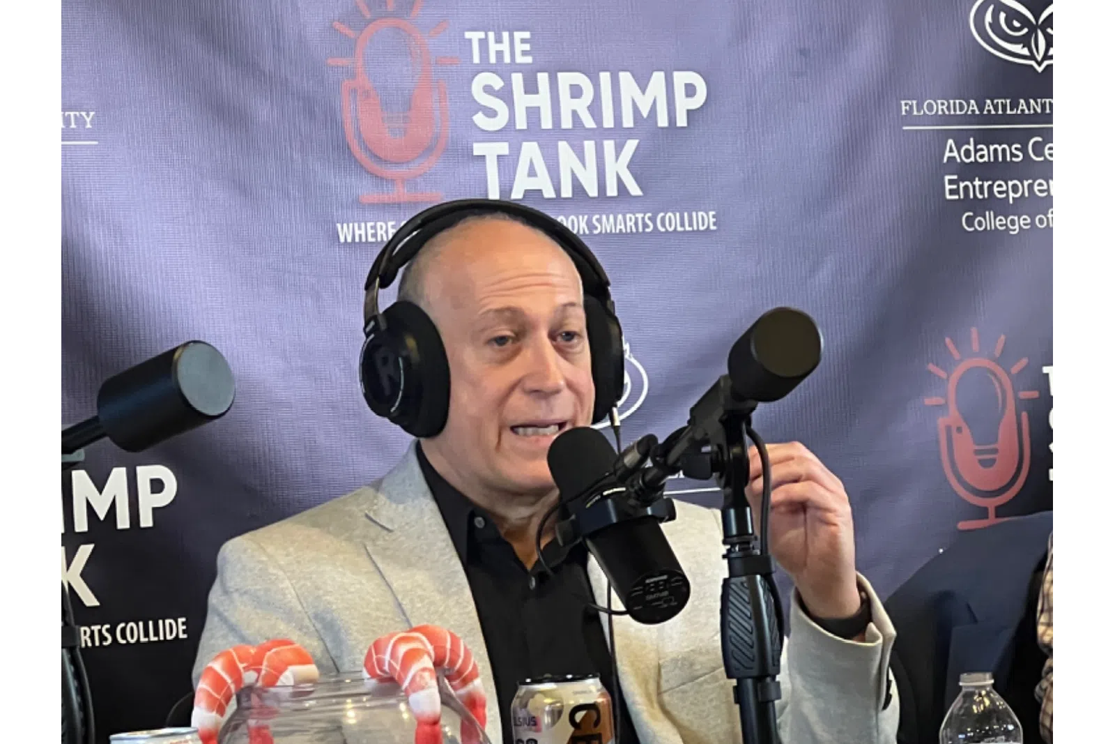 Man wearing headphones speaking into microphone during The Shrimp Tank podcast with shrimp candy jar and Celsius drink cans on desk