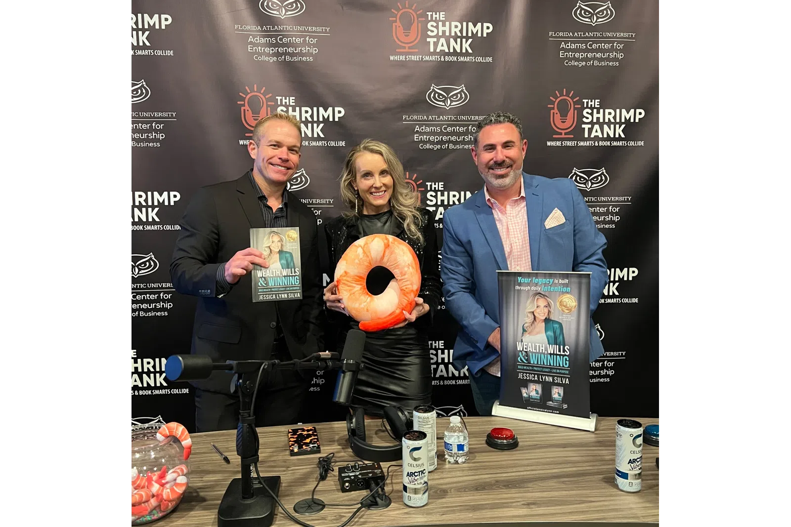 Three people at a podcast table with microphones, holding books and a shrimp-shaped cushion, Shrimp Tank backdrop.