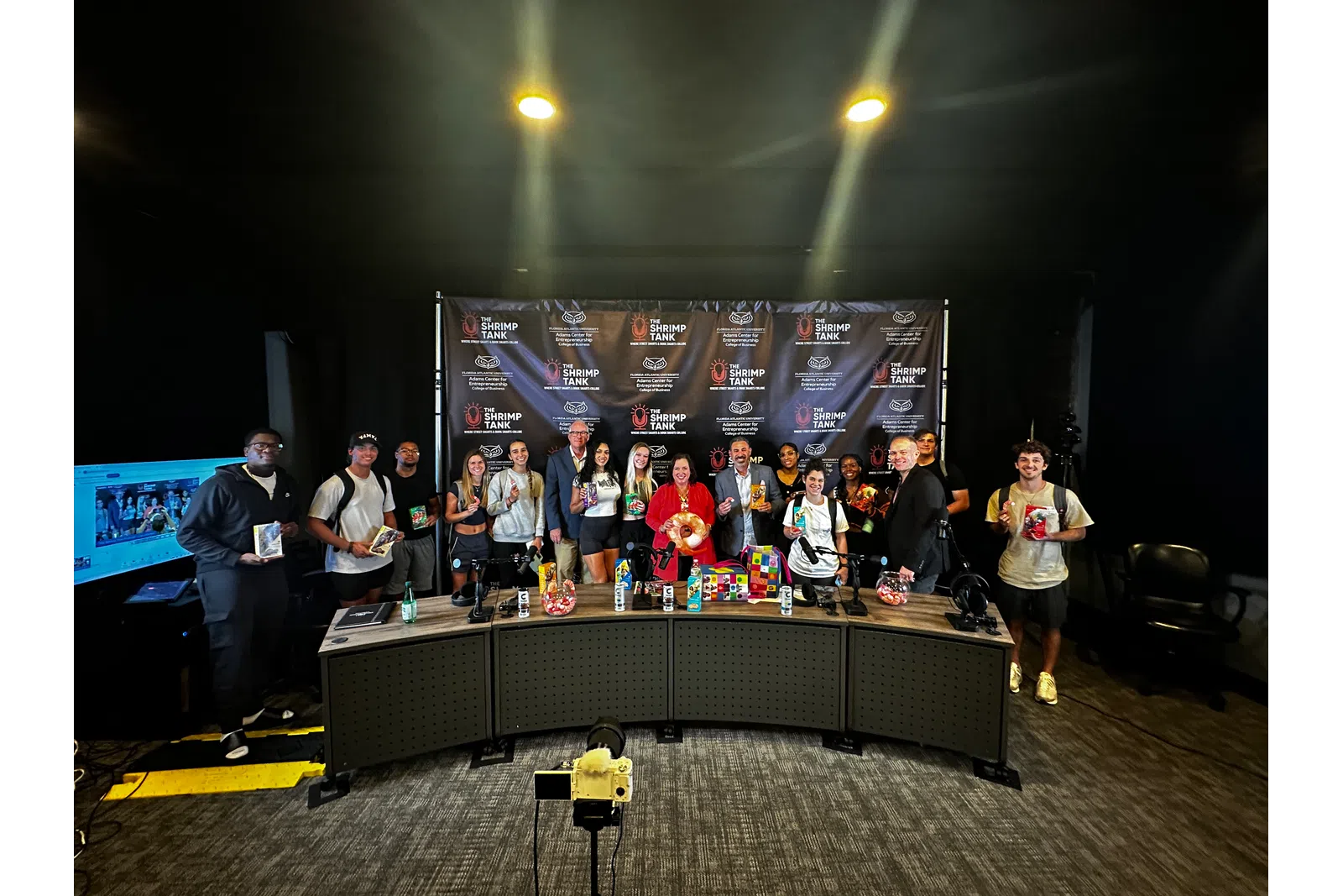 Group photo of attendees at a media event, featuring a branded backdrop and diverse individuals holding merchandise.
