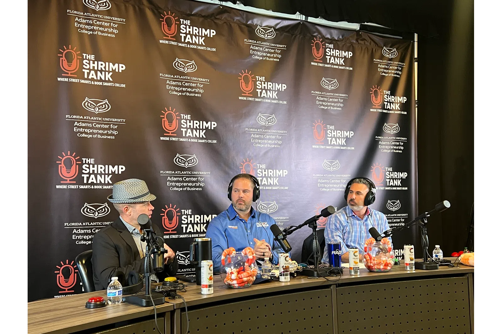 Three men with microphones hosting a podcast in front of The Shrimp Tank backdrop at Florida Atlantic University.