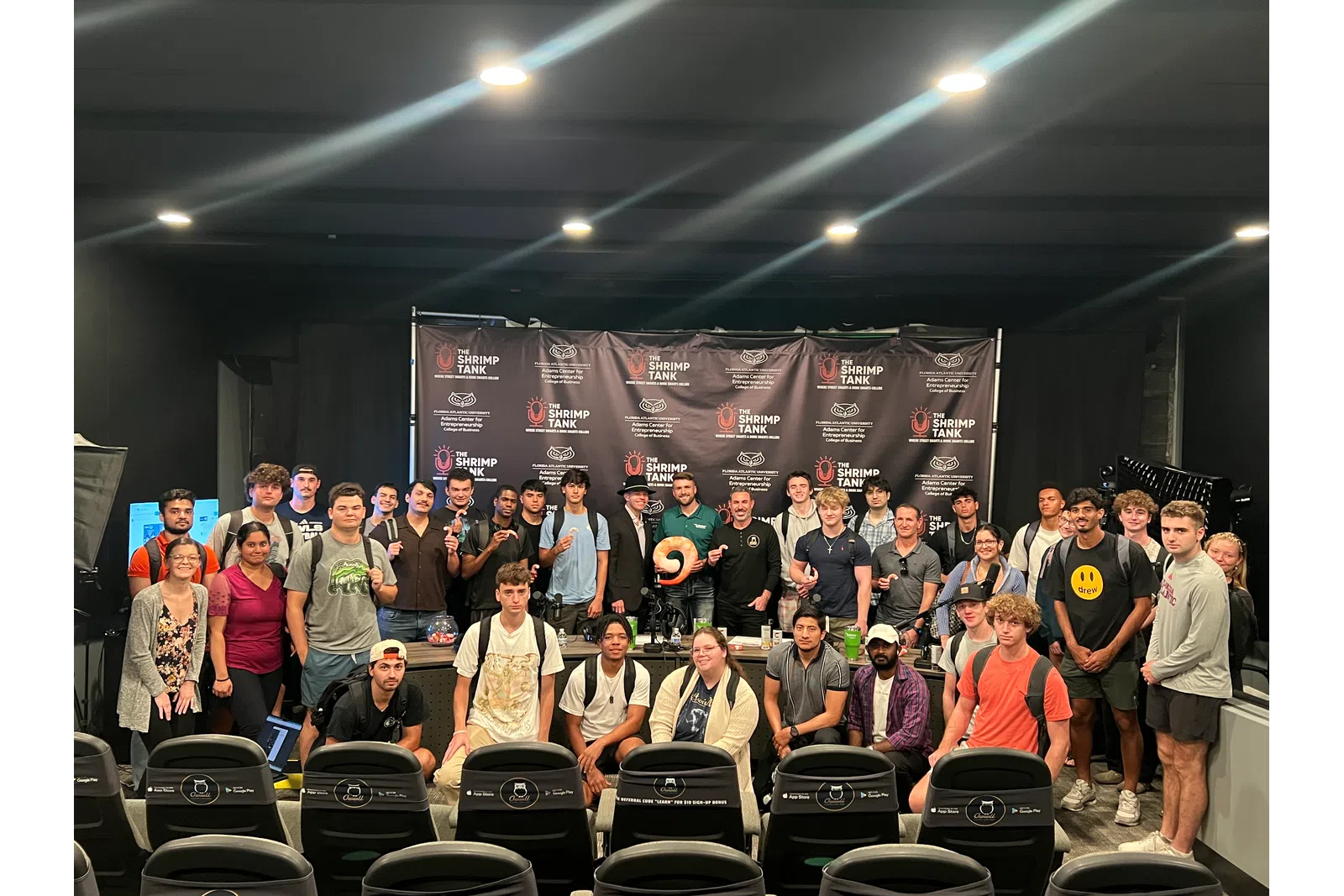 Large diverse group posing in front of a Shrimp Tank banner in an indoor event space with studio lighting.