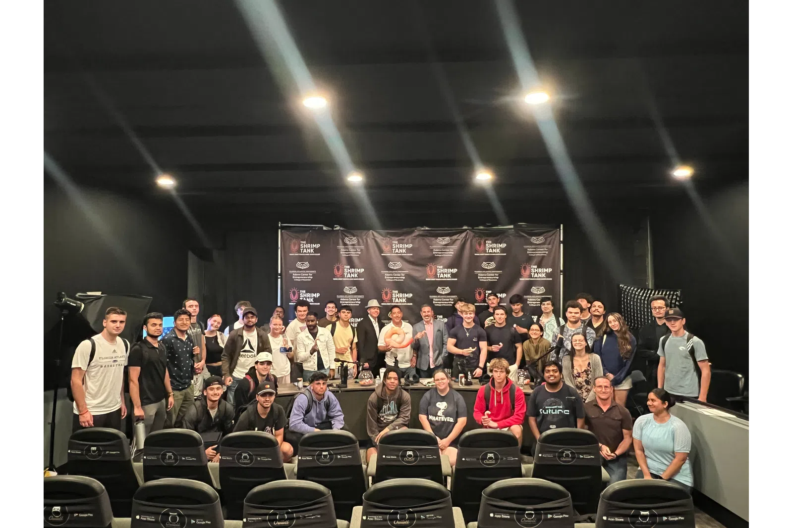 Group photo of diverse people posing in front of a Shrimp Tank event backdrop with studio lighting.