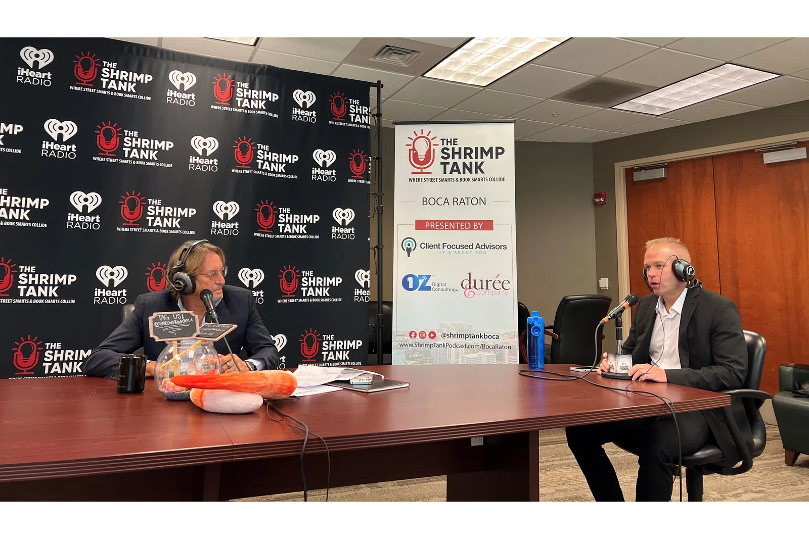 a couple of men sitting at a table with microphones and a sign