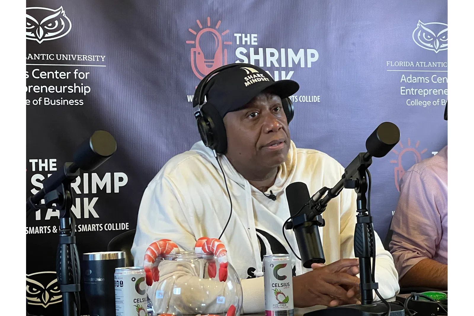 Man wearing headphones and a Shark Mindset cap speaking into a microphone at The Shrimp Tank podcast setup