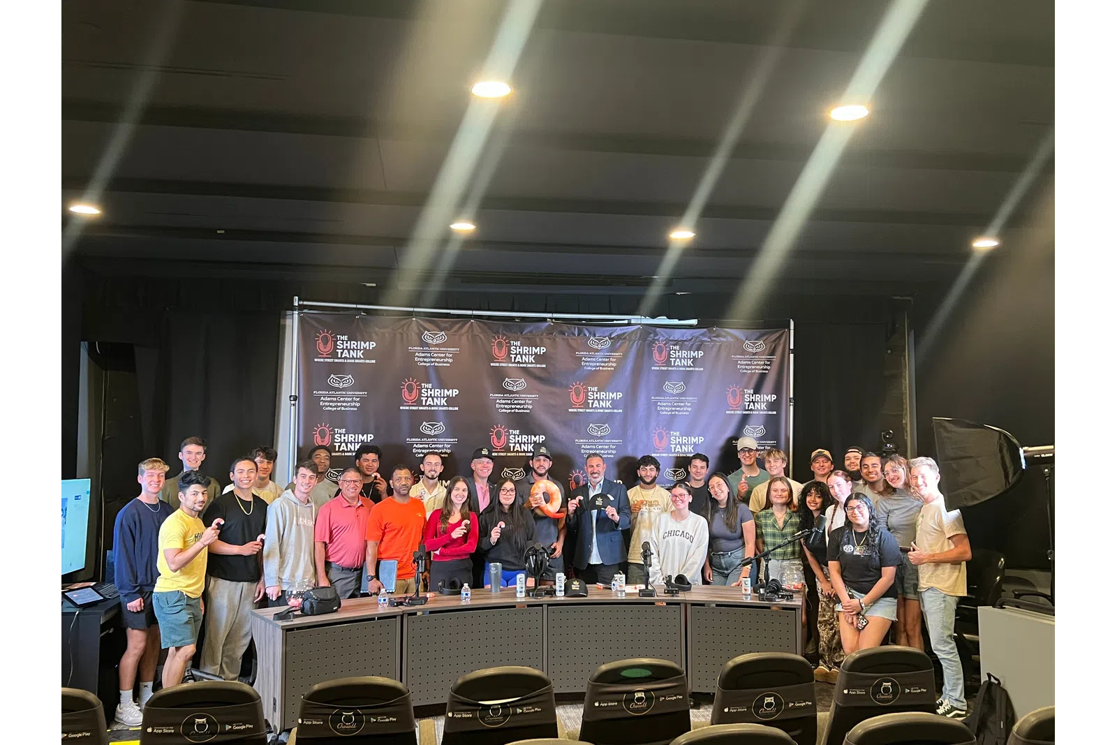 Large group of people posing in front of The Shrimp Tank backdrop with studio lighting and microphones.