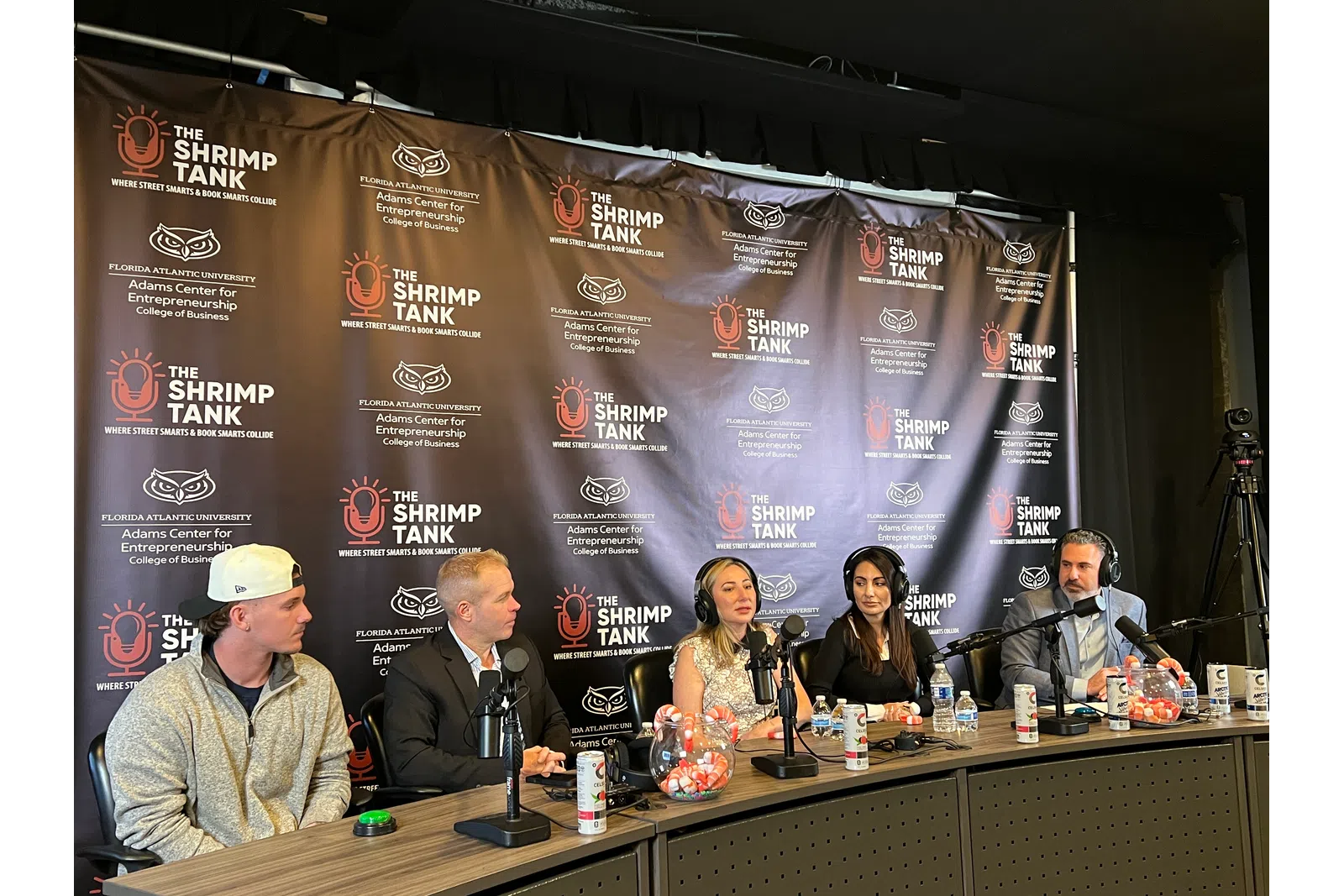 Panel of five speakers with microphones at The Shrimp Tank event backdrop discussing entrepreneurship.