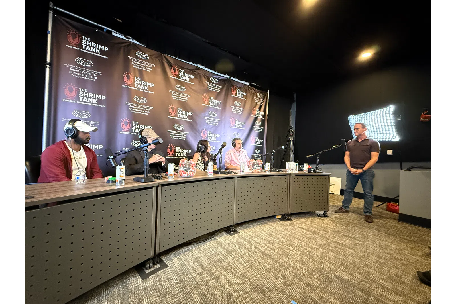 Panel of four judges seated with microphones facing a standing presenter in a podcast studio with The Shrimp Tank backdrop