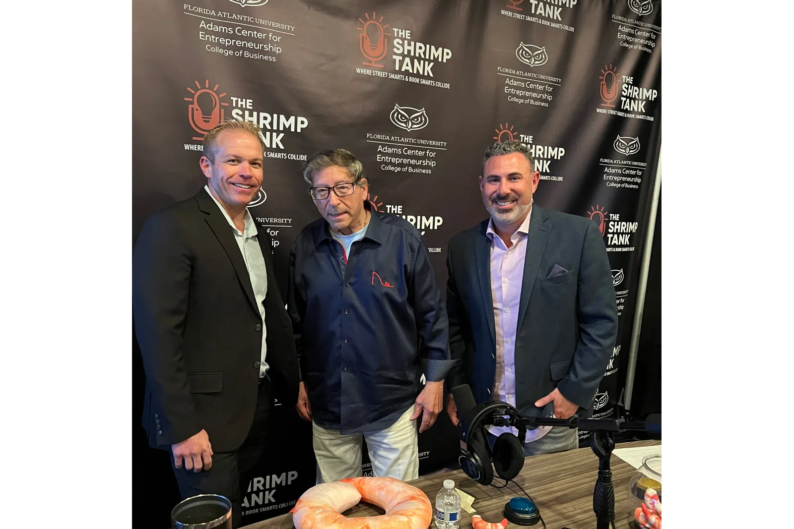 Three men posing in front of The Shrimp Tank podcast backdrop with shrimp-themed props and microphones on a table