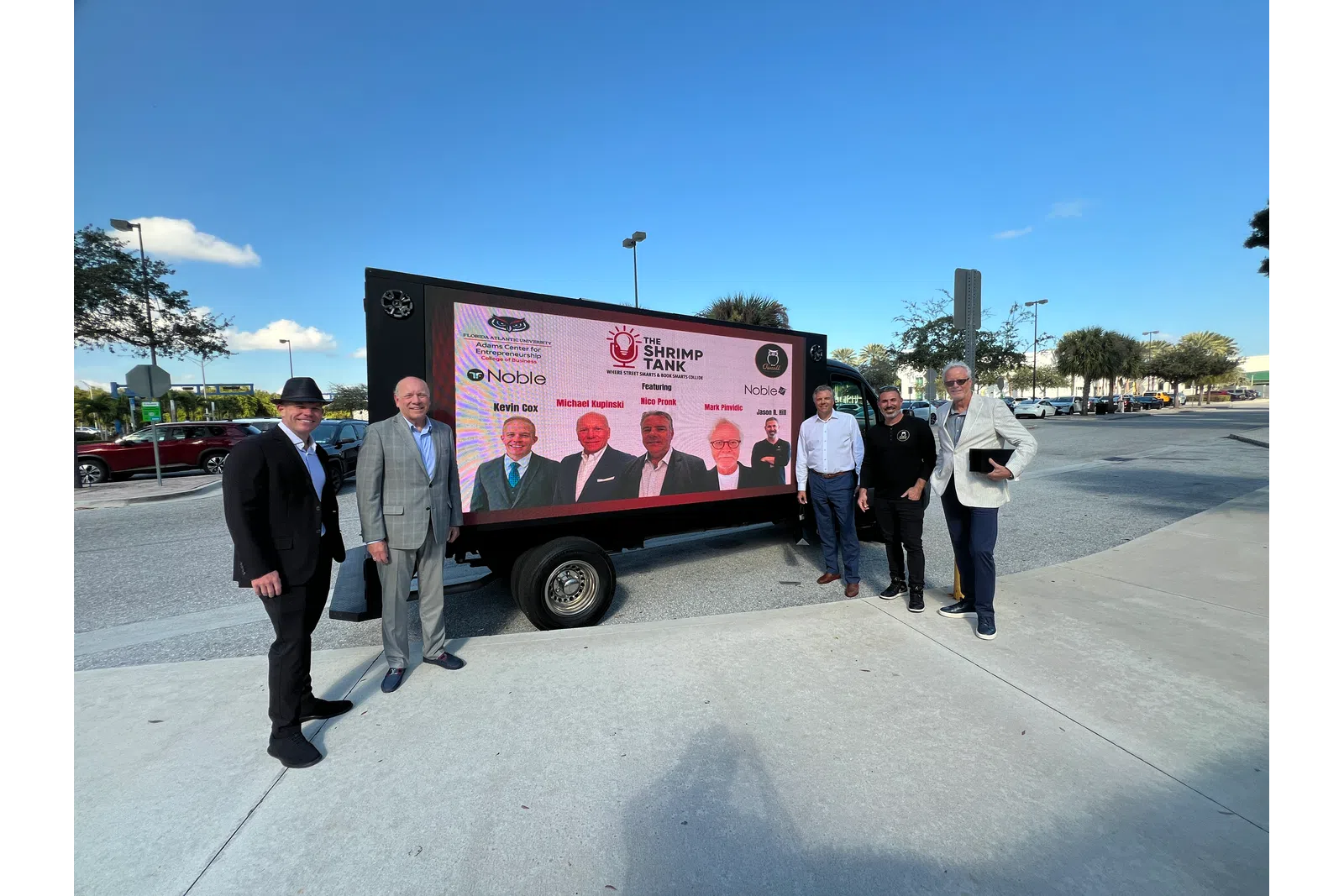 Six men in business attire stand outdoors beside a large digital screen advertising The Shrimp Tank event.