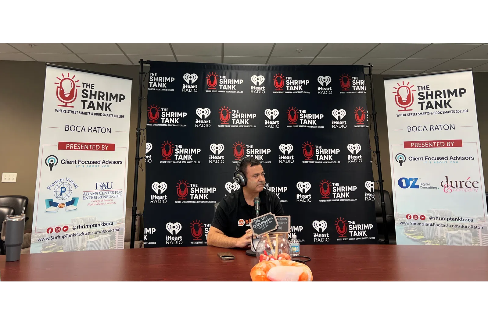 a person sitting at a table with a microphone and a sign behind him