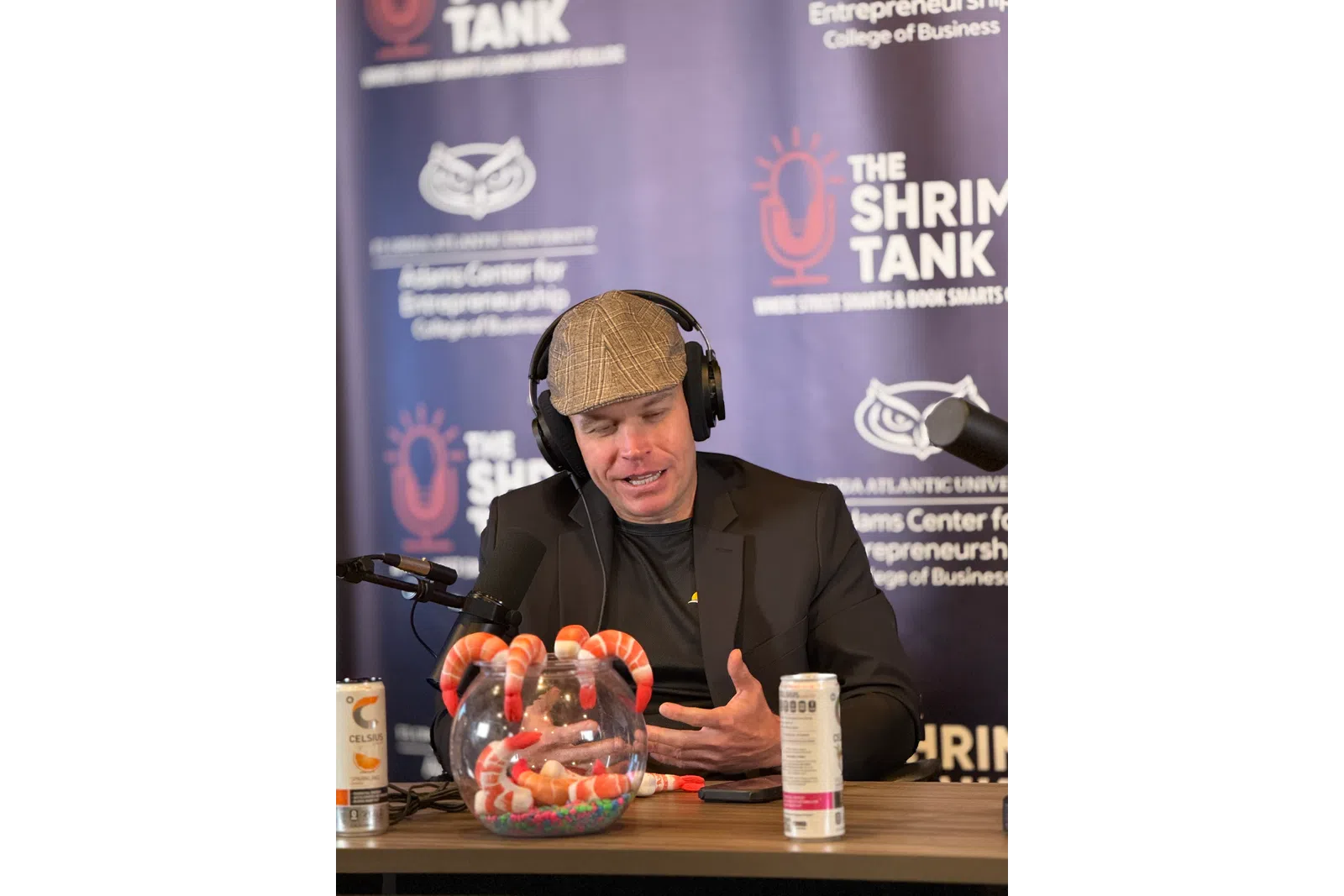Man wearing headphones and a cap speaking into a microphone at a podcast setup with shrimp-themed decorations and energy drinks.