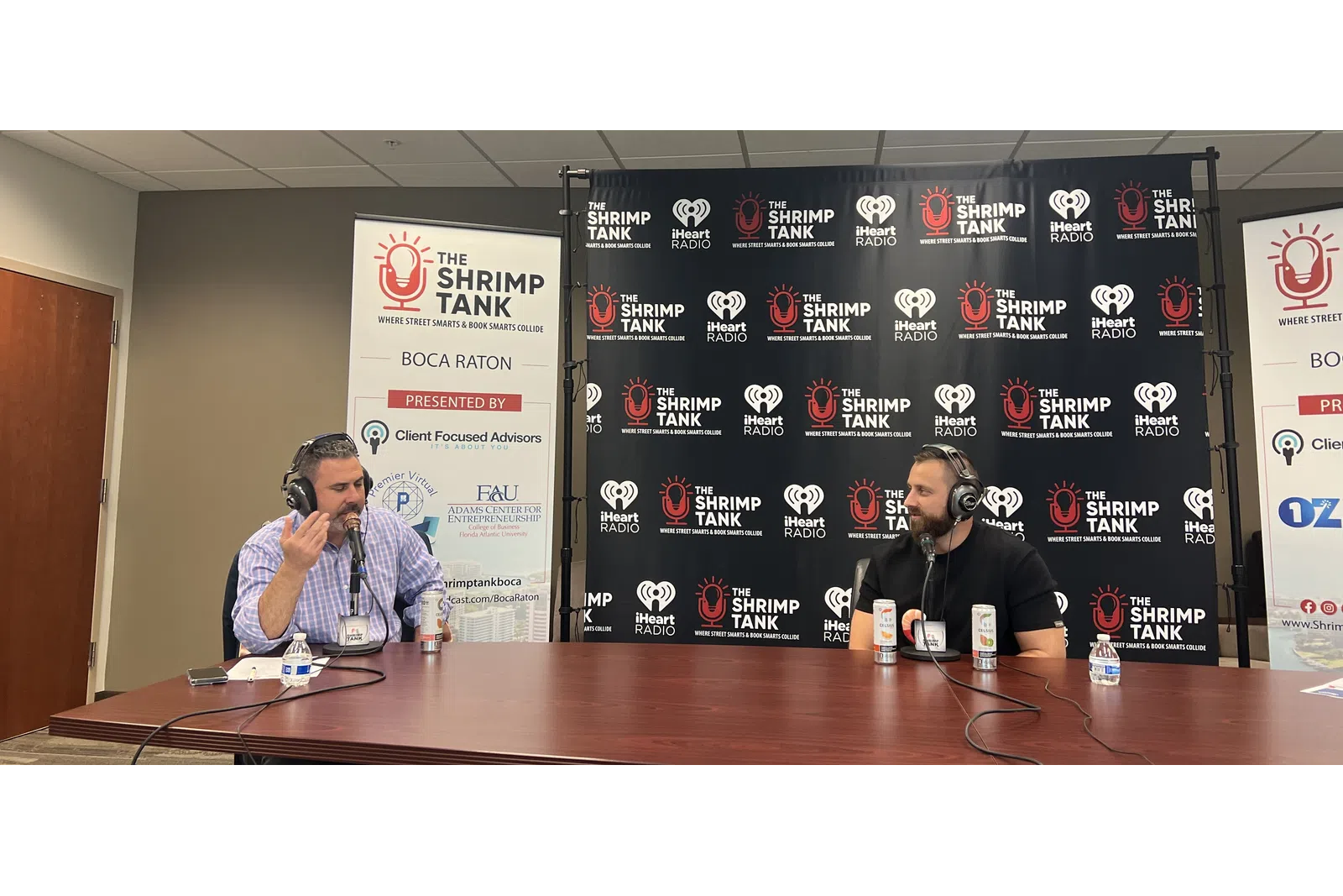 a couple of men sitting at a table with microphones and a sign behind them