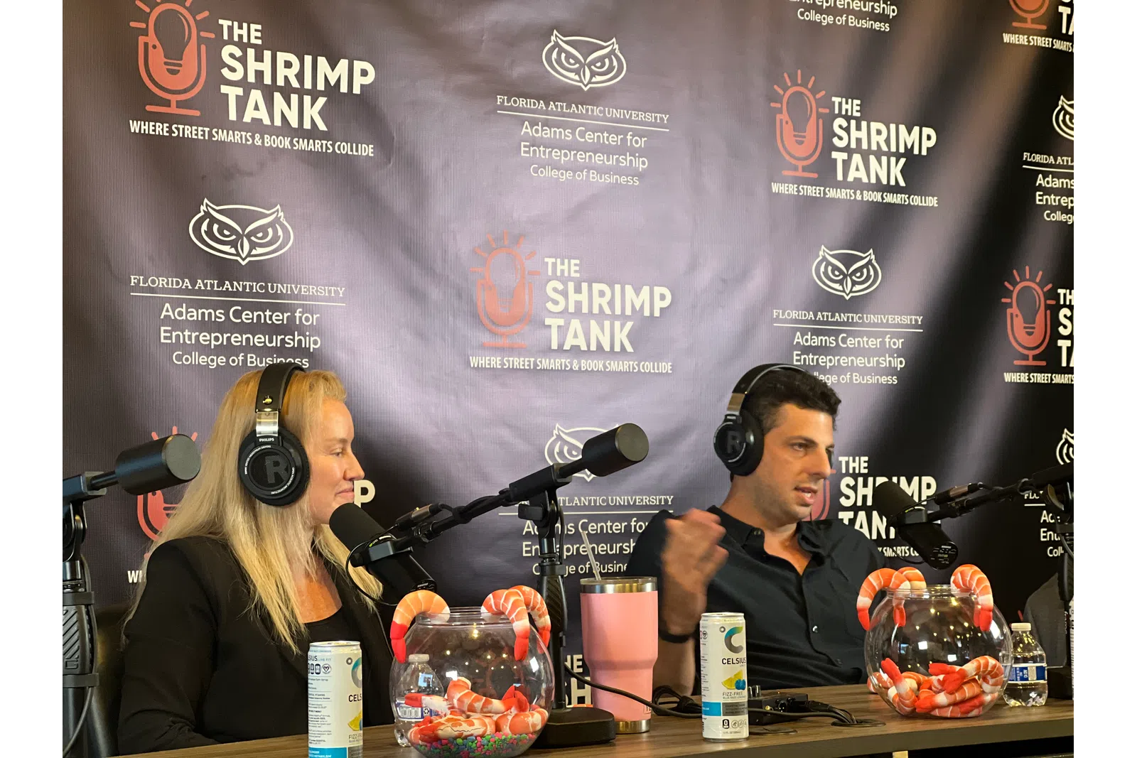 Two podcasters with headphones talk into microphones in front of a backdrop for The Shrimp Tank podcast at Florida Atlantic University.