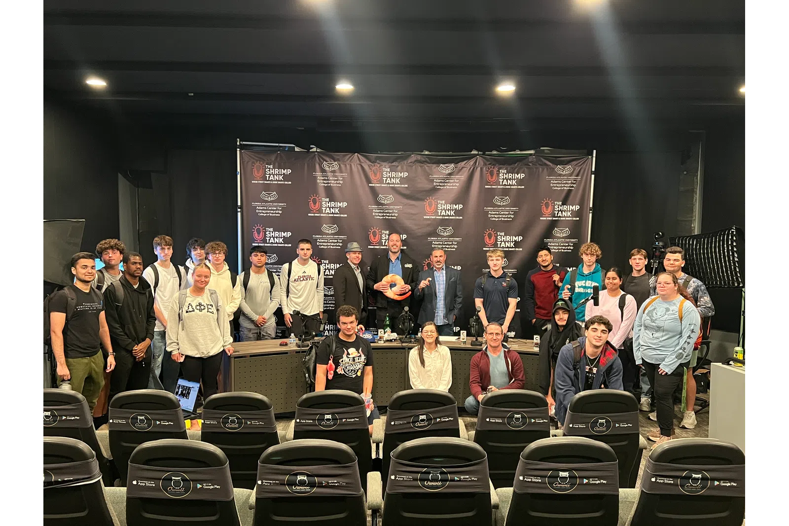 Large diverse group posing in front of The Shrimp Tank branded backdrop in an event space with empty chairs in foreground