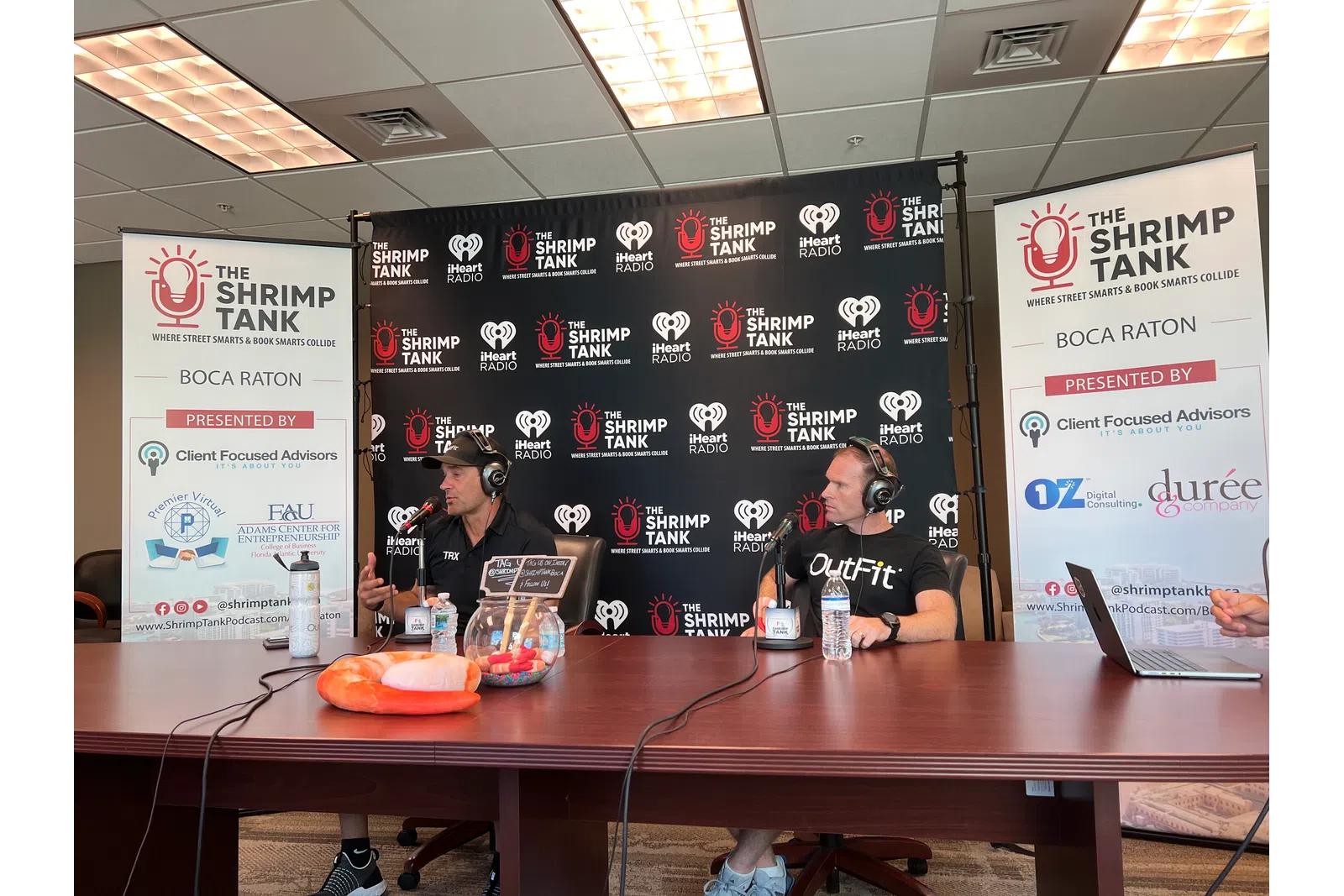 a couple of men sitting at a table with laptops and a sign