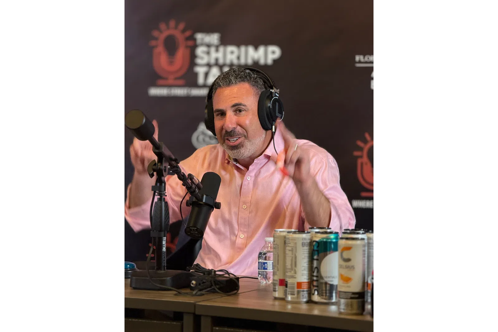 Podcast host wearing headphones speaking into a microphone with energy drinks and water on the table in front.