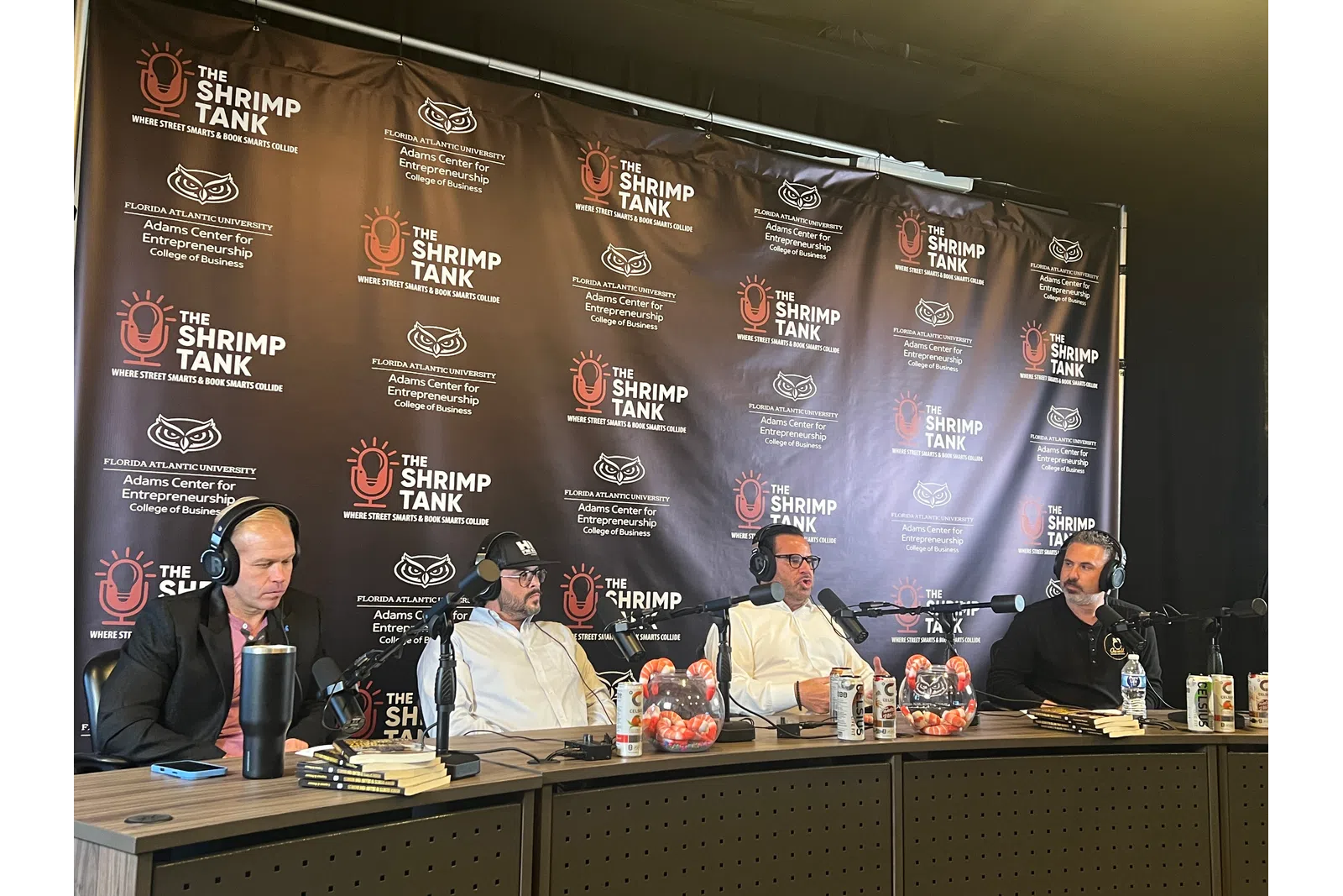 Four men wearing headsets engaged in a podcast discussion in front of The Shrimp Tank banner at Florida Atlantic University.