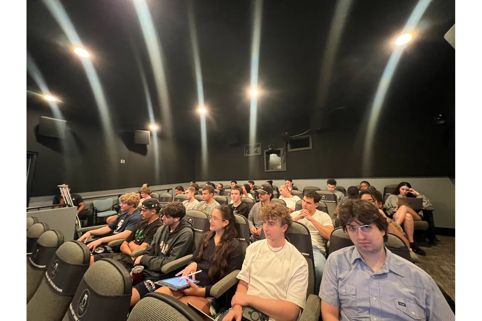 Group of students attentively seated in a dark lecture hall with black walls and ceiling lights.
