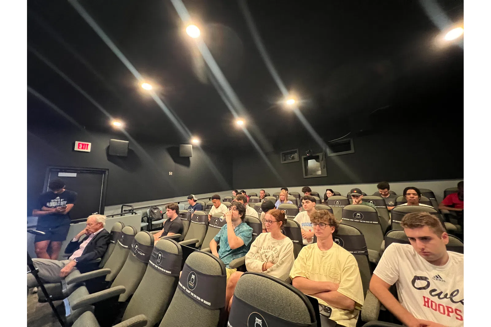 Group of people seated in a small, dimly lit theater or lecture room with black walls and ceiling lights.