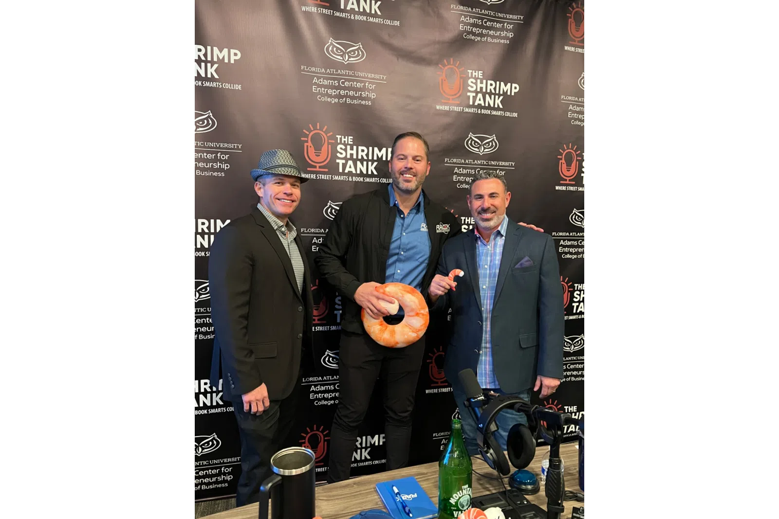 Three men posing at The Shrimp Tank event with branded backdrop, shrimp-shaped item, and recording equipment.