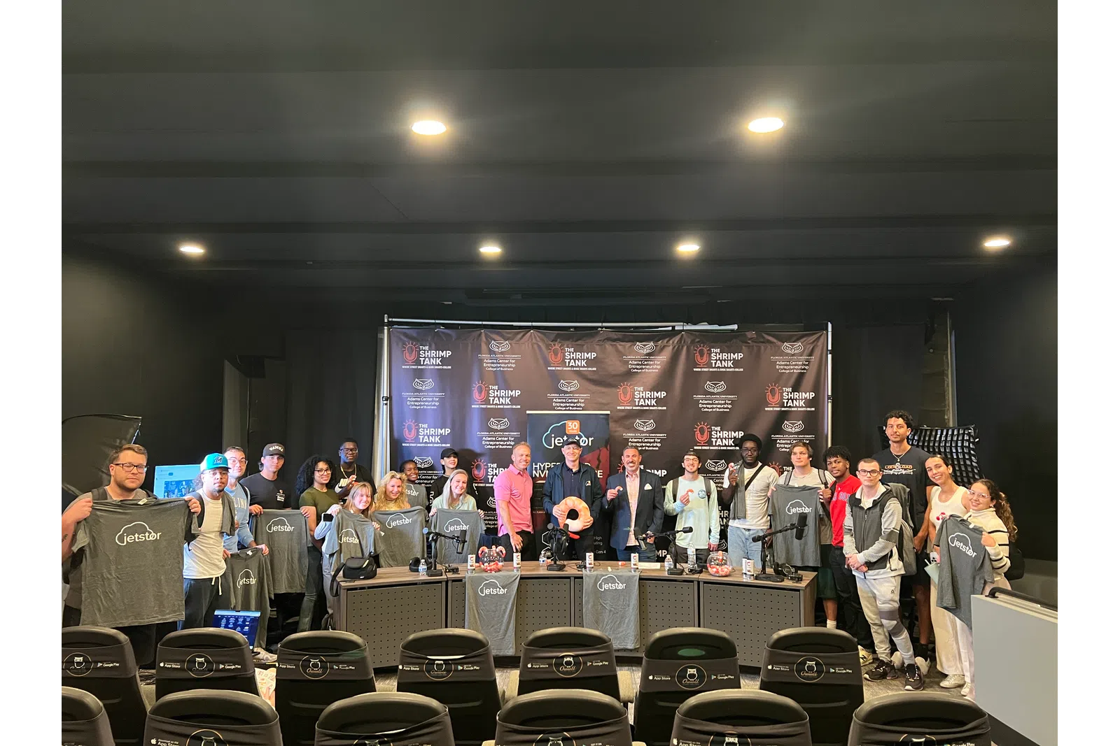 Group photo of attendees wearing branded shirts at a promotional event with a backdrop.