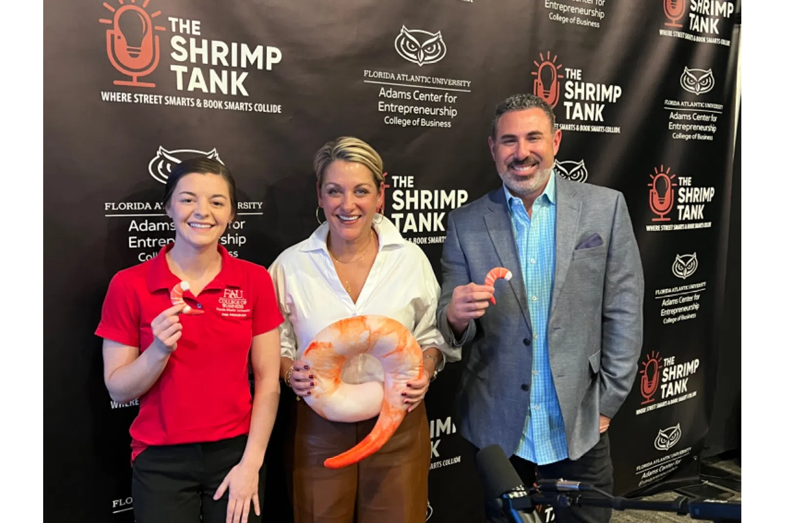 Three people posing in front of The Shrimp Tank backdrop holding large shrimp props in a podcast studio setting