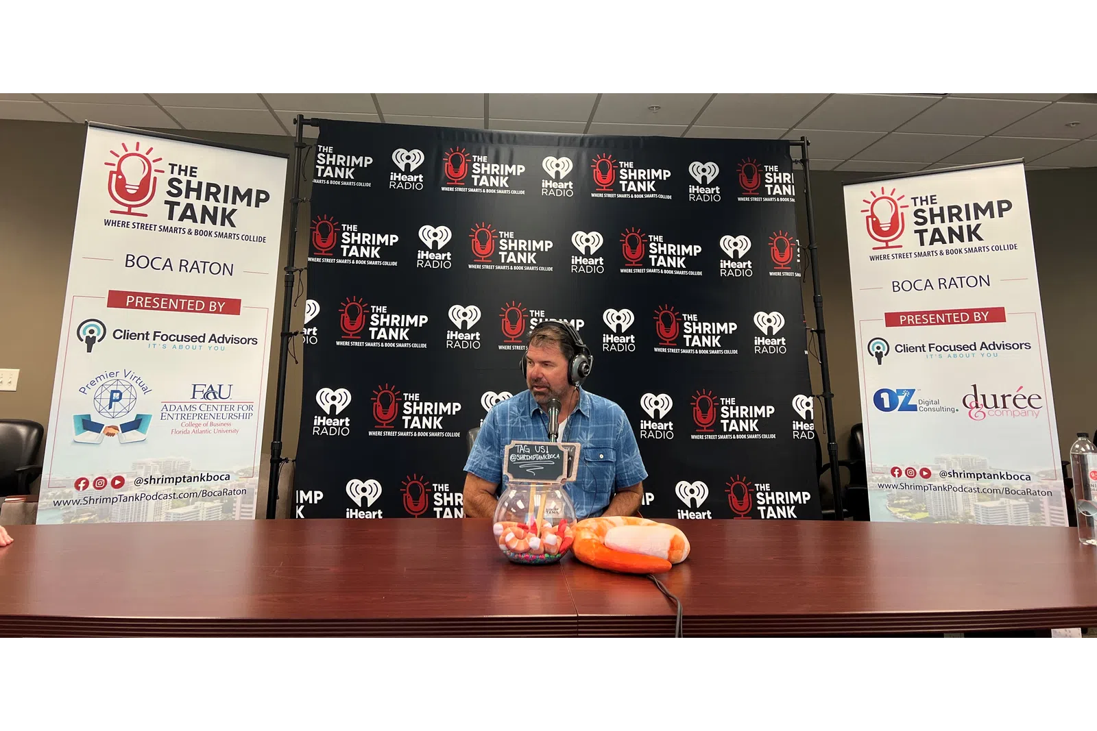 a person sitting at a table with a microphone and a sign behind him