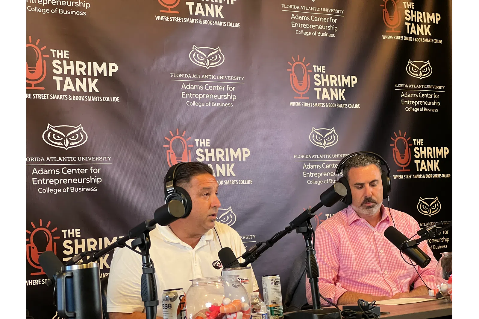 Two men wearing headphones speaking into microphones during The Shrimp Tank podcast recording with branded backdrop