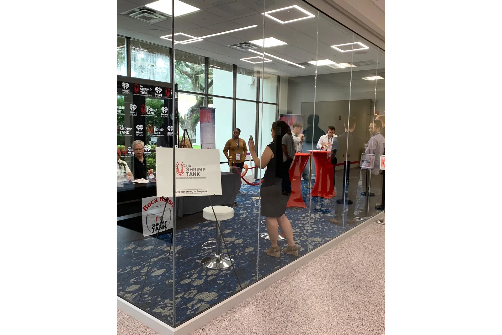 People attending a live recording of The Shrimp Tank podcast in a glass-walled conference room with banners and tables.